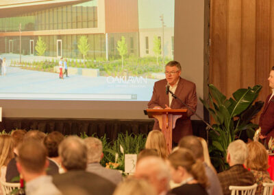 A man stands at a podium speaking to a seated audience, with a large screen behind him displaying an architectural rendering labeled OAKLAWN HOT SPRINGS, ARKANSAS. Another person stands beside him.
