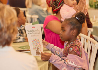 A young girl sits at a table, reading a brochure titled Rooted in Reading, Growing for Good. Other children and adults are seated around her, and the setting appears to be a community or school event.