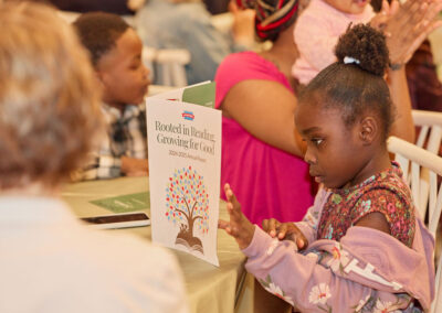 A young girl sits at a table looking at her hand, with a report titled Rooted in Reading, Growing for Good propped up in front of her. Other children and adults are seated nearby in a casual indoor setting.