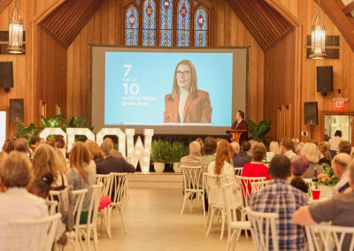 A large audience sits in a wooden hall watching a presentation. A woman on a screen displays the text “7 out of 10 reading below grade level.” Large white letters spelling “GROW” are on stage with plants.