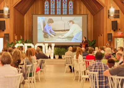A crowd seated at round tables in a large wooden hall watches a speaker at a podium. A projector screen behind shows two people playing with toys on the floor. Large letters spelling GROW are illuminated onstage.