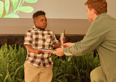 A young boy in a plaid shirt receives a trophy and books from an adult on stage. Behind them, a screen displays the text Favorite things to do outside of school: Building Legos. Lush green plants line the stage.