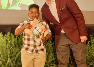 A young boy holding a trophy and a colorful certificate stands next to a smiling man in a maroon blazer, both posing in front of green plants and a screen that mentions “Building Legos.”.