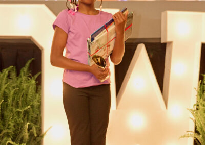 A young girl in a purple shirt and black pants stands on stage holding a trophy and gifts, smiling. Large illuminated letters and green plants decorate the stage behind her.