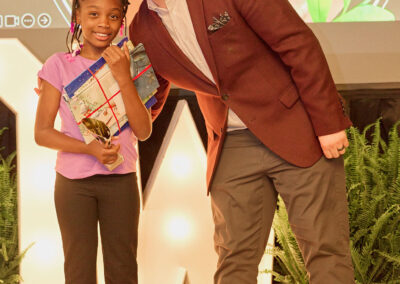 A smiling man in a maroon blazer stands next to a young girl in a purple shirt holding a gift and a certificate. They pose together on stage in front of a screen showing a childs photo. Green plants decorate the stage.