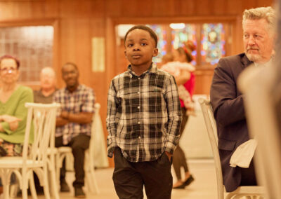 A young boy in a plaid shirt and black pants walks confidently down an aisle in a warmly lit room, while seated adults watch in the background.