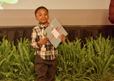A smiling young boy stands indoors holding books and a ribbon, in front of a row of potted plants. Behind him, a screen reads, Favorite things to do outside of school: Play outside with his sister.