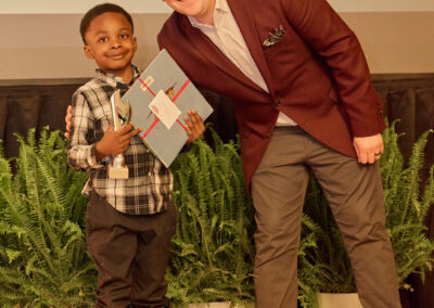 A young boy holding a trophy and a certificate stands next to a smiling man in a maroon blazer. They pose in front of green plants. Text in the background reads, Play outside with his sister.