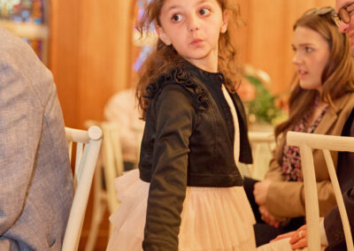 A young girl in a frilly cream dress and black jacket stands beside a seated group in a warmly-lit room with wooden walls and stained glass windows, looking over her shoulder.