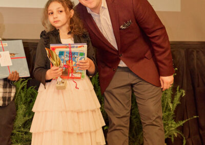 A young girl in a cream dress holds awards and poses next to a smiling man in a maroon blazer. They stand in front of a stage with a projection that says “Jumping on the trampoline” in the background.