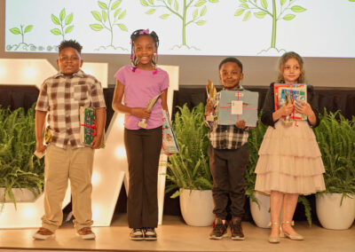 Four young children stand side by side on a stage, smiling and holding trophies and books, with potted plants and a backdrop featuring illustrations of green leafy plants behind them.