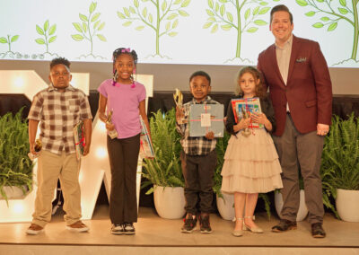 Four young children stand on stage holding prizes and trophies, smiling next to an adult man in a maroon blazer. Potted plants and a backdrop with illustrated trees are behind them.