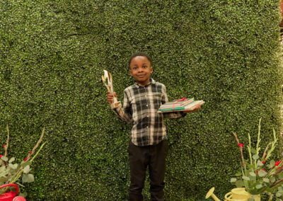 A young boy stands in front of a green wall with a neon AR KIDS READ sign, holding a trophy in one hand and a stack of books in the other. There are plants and watering cans on either side of him.