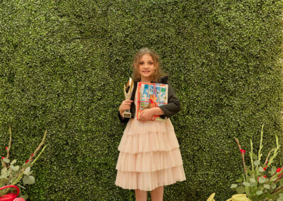 A young girl in a pale pink dress stands smiling in front of a green leafy backdrop with a neon sign that reads AR KIDS READ. She holds a trophy and a book, with colorful watering cans and plants on either side of her.