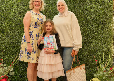 Three women—one young girl in a pink dress holding a book, an older woman in a floral dress, and a woman in a hijab and sweater—stand smiling in front of a green, leafy backdrop with a neon sign reading AR KIDS READ.