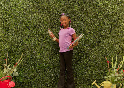 A young girl in a purple shirt stands smiling in front of a green leafy wall with a neon sign reading AR Kids Read. She holds books and pencils, with two watering cans and plants on either side of her.