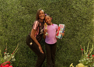 A woman and a girl pose and smile in front of a green leafy wall with a neon sign that says AR KIDS READ. The girl holds a book and a trophy; watering cans and plants are on the floor nearby.