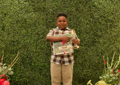 A young boy stands smiling, holding a trophy and a colorful sign, in front of a green leafy backdrop with a neon AR KIDS READ sign. Decorative plants and watering cans are on the floor beside him.