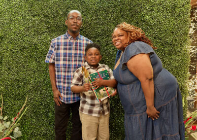 A smiling family of three poses in front of a green hedge wall with a neon sign reading “AR KIDS READ.” The child in the center holds books, while the adults stand on either side, all dressed in casual attire.
