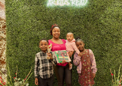 A woman stands with three children in front of a green hedge wall with a neon “AR KIDS READ” sign. The children hold books and a trophy. Decorative plants with watering cans are on each side of the group.