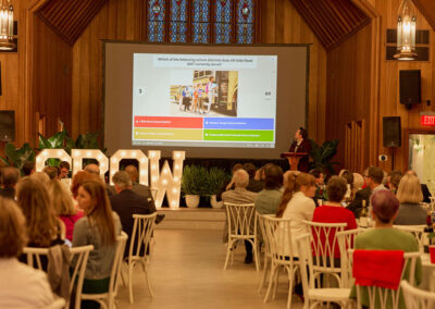 A group of people sit at tables in a large hall with wood paneled walls, watching a presentation projected on a screen. A speaker stands at a podium, and illuminated letters spell “GROW” at the front of the room.