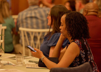 A woman sits at a table in a busy indoor event, looking at her phone. She is wearing a floral dress and has short curly hair. Other people are seated nearby, and papers and a plastic cup are on the table.