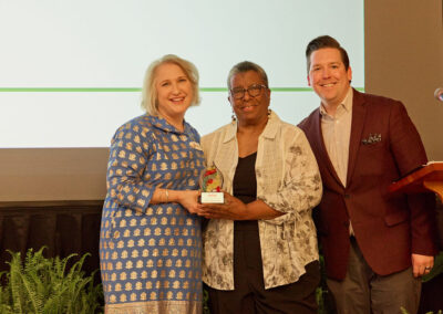 Three people stand together on a stage, smiling at the camera. The woman in the center holds a small award. Behind them, a screen reads Tutor of the Year Nancy Robinson Lott. There are potted ferns in the foreground.