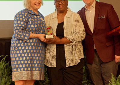 Three people stand together smiling; the woman in the center holds an award. They are posing in front of greenery and a presentation screen displaying text.