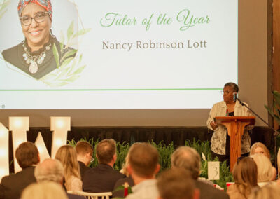 A woman stands at a podium addressing an audience at an indoor event. Behind her, a large screen displays Tutor of the Year, Nancy Robinson Lott with her photo and decorative greenery.