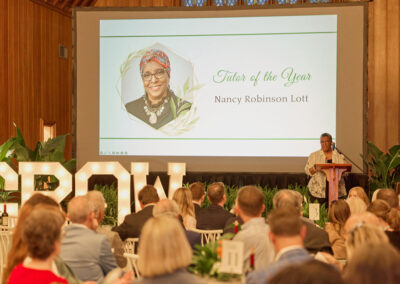 A woman stands at a podium speaking to an audience in a wooden hall. Behind her, a large screen displays Tutor of the Year, Nancy Robinson Lott with her photo and greenery-themed graphics.