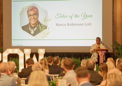 A woman stands at a podium addressing an audience; behind her is a large screen showing her photo and the text “Tutor of the Year Nancy Robinson Lott.”.