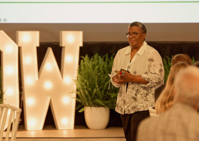 A woman stands and speaks in front of a large illuminated W and green plants, addressing an audience at an indoor event. She holds an object and wears glasses and a patterned shirt.