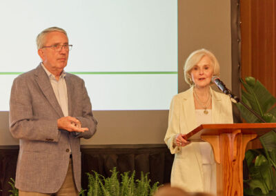 An older woman speaks at a wooden podium with a microphone, while an older man stands beside her. Both are dressed in light-colored suits, and there are green plants and a large screen in the background.
