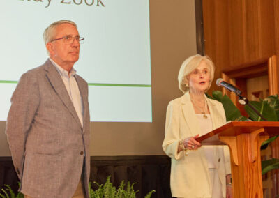 A man in a gray blazer and a woman in a white outfit stand at a podium. The woman speaks into a microphone. Behind them is a projection screen and a potted plant is visible beside the podium.