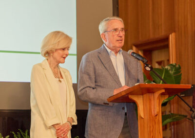 An older man in a blue blazer speaks at a podium with a microphone, while an older woman in a white outfit stands beside him. They are indoors, in front of a screen and wooden wall, with a plant nearby.