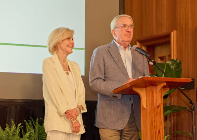An older man speaks at a wooden podium with a microphone, while an older woman stands beside him smiling. They are indoors near a projection screen and plants, both dressed in light-colored, formal clothing.