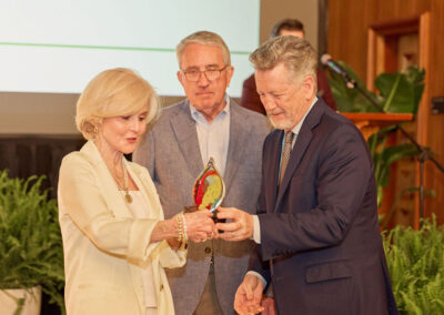 Three older adults, two men and one woman, stand together indoors. The woman hands a colorful award to one man while the other man looks on. Fern plants and a microphone are visible in the background.