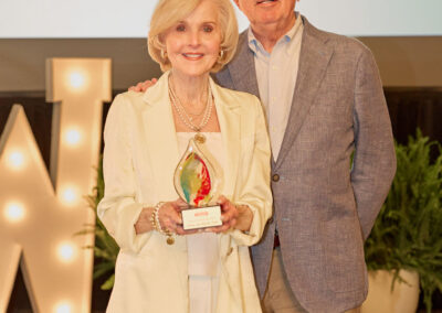 An older woman in a cream suit holds a colorful glass award and stands next to an older man in a blue blazer, both smiling at an indoor event with a lit “W” and greenery in the background.