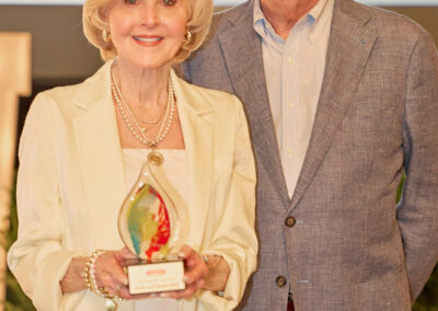 An older woman holding an award stands next to an older man. Both are dressed in formal attire and smiling at the camera. The background shows a blurred stage setting with greenery and a projection screen.