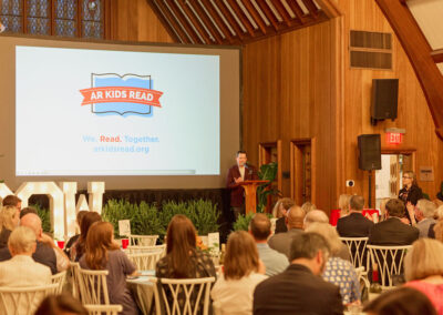 A speaker stands at a podium addressing a seated audience in a wood-paneled room, with a large screen behind him displaying the AR Kids Read logo and the phrase We. Read. Together.