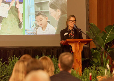 A woman stands at a wooden podium speaking into a microphone in front of an audience. Behind her, a screen displays images of children and an adult in a classroom setting. Green plants decorate the stage.