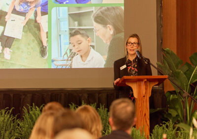 A woman stands at a podium speaking to an audience, with a large screen behind her displaying images of children and a teacher in a classroom setting. Green plants decorate the stage.