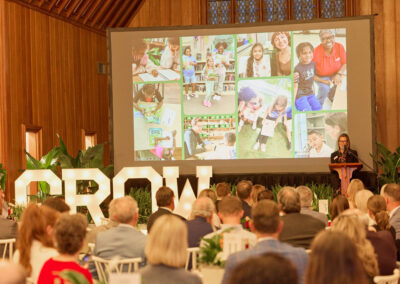 A woman speaks at a podium to an audience in a wooden hall. Behind her, a large screen displays photos of children reading and learning. Large illuminated letters spell GROW in front of the stage, with greenery around the room.