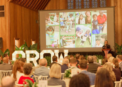 A woman speaks at a podium to a seated audience in a large room. Behind her, a screen displays a collage of children and adults reading. Large illuminated letters spell GROW near the stage.