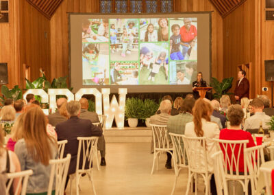 A woman speaks at a podium in front of an audience in a large hall. Behind her is a screen displaying children’s photos and large illuminated letters spelling GROW, surrounded by plants.