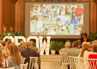 A formal event in a large wooden hall with people seated at tables, facing a stage. A speaker stands at a podium beside a large screen displaying a collage of children and activities. Large white letters spell GROW on stage.