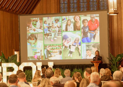 A speaker stands at a podium in a wood-paneled hall, addressing an audience seated at tables. Behind her is a large screen displaying a collage of children engaged in educational activities.