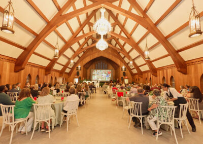 A large group of people sits at round tables in an elegant hall with wooden beams, chandeliers, and high arched ceilings, watching a presentation on screens at the front of the room.