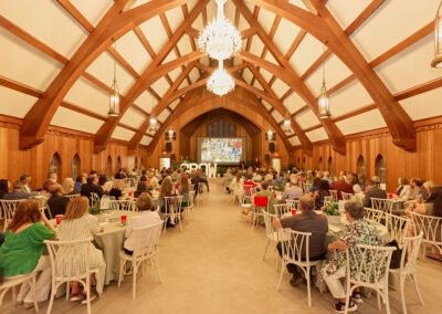 A large group of people sits at round tables in a wood-paneled banquet hall with vaulted ceilings and chandeliers, facing a stage with a large screen displaying colorful images.