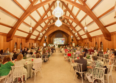 A large group of people sit at round tables in a spacious, wood-paneled hall with vaulted ceilings, chandeliers, and arched windows, watching a presentation on a large screen at the front.